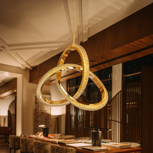 A luxurious modern living room featuring a gold chandelier made of three interlocking rings hanging from the ceiling. The space includes a brown leather sectional sofa, a black round coffee table, and white accent chairs, all illuminated by warm natural and artificial light that highlights the chandelier’s golden texture and the room’s contemporary deco