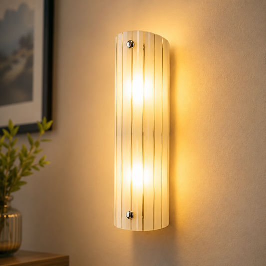 Wall-mounted cylindrical light fixture on a neutral wall with a plant and framed picture in the background.