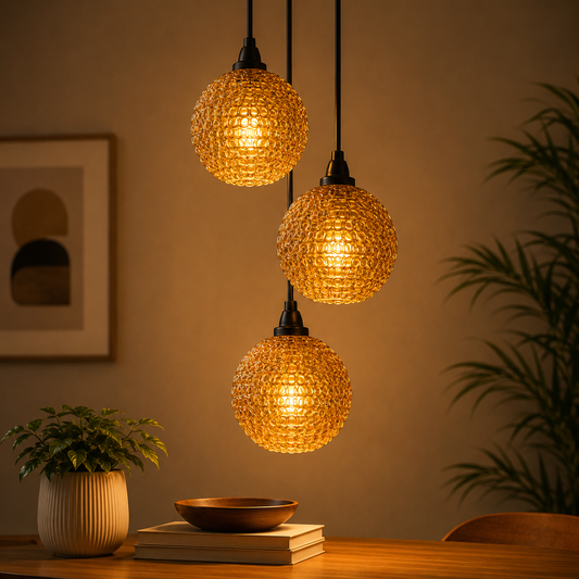Three glowing pendant lights hanging above a table with a plant and books in a warm-toned room.